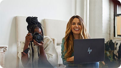 Deux personnes : l'une tient un appareil photo dans ses mains, l'objectif pointé devant elle, et la seconde tient un ordinateur portable en souriant.